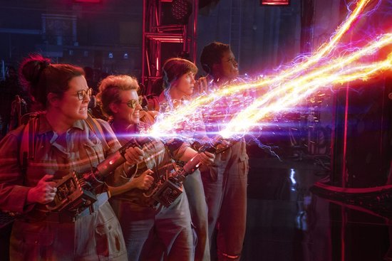 Vier vrouwen in Ghostbusters-uniformen die tegelijkertijd met protonenpakken op iets schieten.