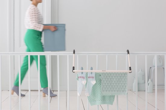 Wit droogrekje met grijze ophanghaken, hangend aan een lichte balustrade. Een gebreide trui en sokken drogen erop. Een vrouw draagt een mand.