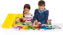 Een jongen en een meisje spelen met LEGO, omringd door een grote gele LEGO-opbergdoos en een berg kleurrijke stenen.