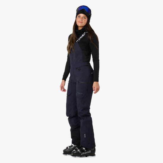 Donkerblauwe skibroek met bretels, gedragen door een model met skiuitrusting.