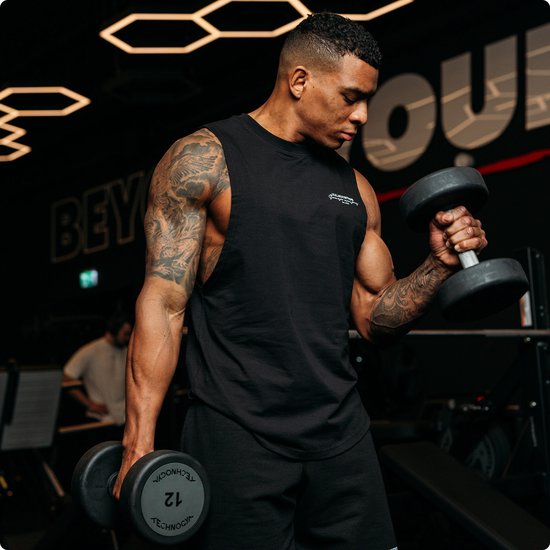 Man met getatoeëerde armen in een zwarte tanktop die dumbbells heft in een sportschool.