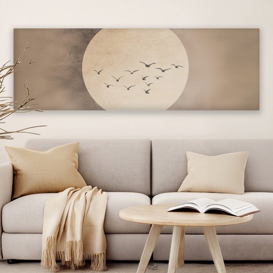 Een langwerpig canvas met een grote, licht beige cirkel tegen een beige achtergrond. Een groepje donkere vogels vliegt in formatie voor de cirkel. Hangt boven een lichte bank met kussens en een plaid.