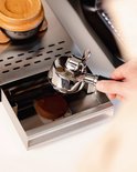 Een hand houdt een zilverkleurige portafilter met een bruin houten handvat boven een uitklaplade van geborsteld staal.