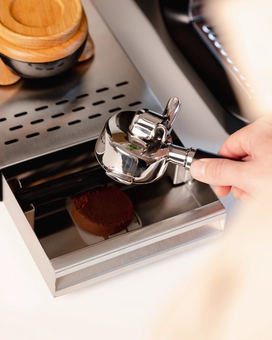 Een hand houdt een zilverkleurige portafilter met een bruin houten handvat boven een uitklaplade van geborsteld staal.