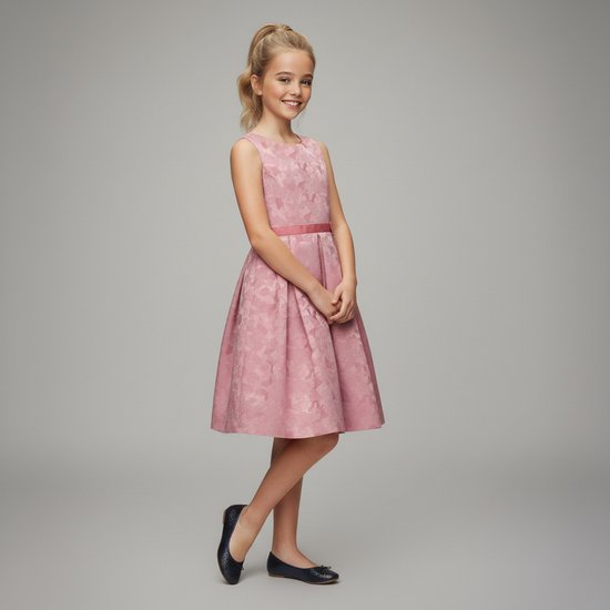 Model draagt een lichtroze jurk met strikdetail en bloemenprint. Ze draagt er zwarte schoenen bij.