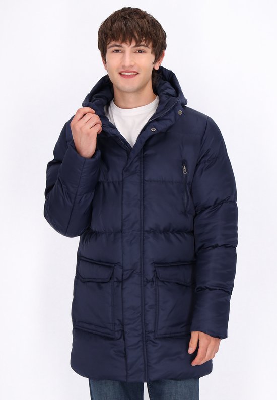 Jonge man met donkerblauwe MO parka met capuchon, staand met handen aan de capuchon en zakken. Draagt spijkerbroek.