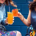 Deux femmes trinquent avec des verres Duralex Manhattan contenant des boissons orangées, devant un mur bleu.