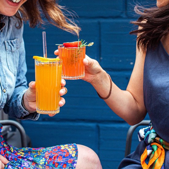 Deux femmes trinquent avec des verres Duralex Manhattan contenant des boissons orangées, devant un mur bleu.