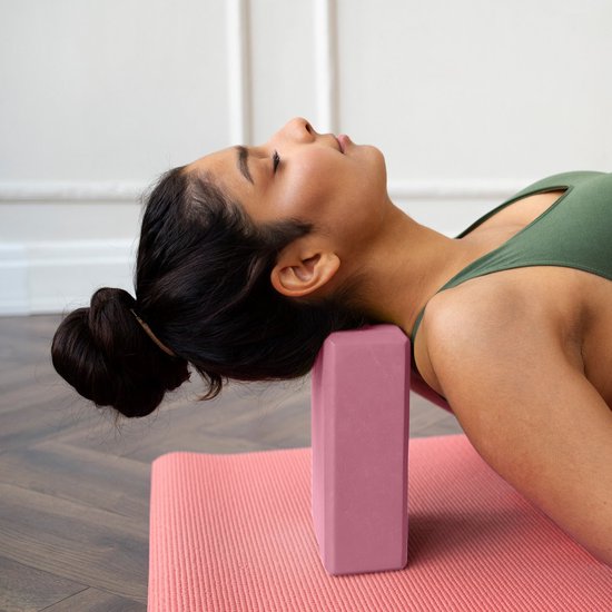 Vrouw in groene sportkleding met hoofd op roze yogablok, liggend op een roze mat.