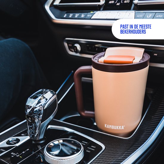 Beige Kambukka tumbler met bruin handvat in een auto bekerhouder. De tekst 'Past in de meeste bekerhouders' is zichtbaar.