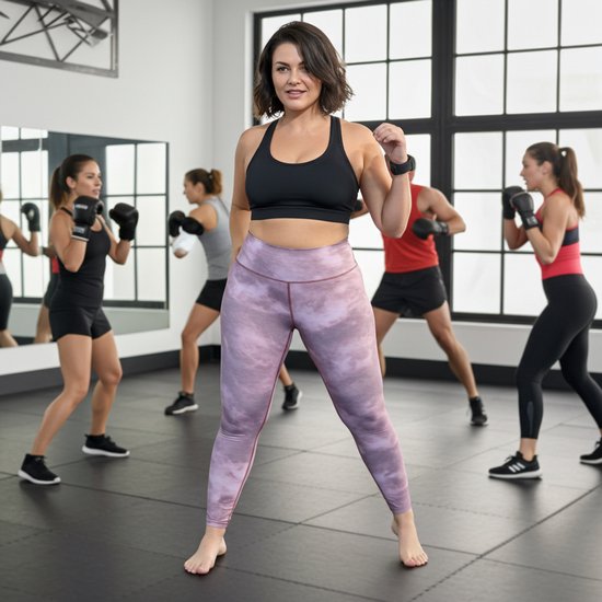 Vrouw met roze tie-dye legging en zwarte sportbeha in een fitnessstudio, met bokstrainers op de achtergrond.