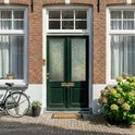 Voordeur van baksteen met groene deuren en ramen met mozaïek raamfolie. Fiets en planten buiten.