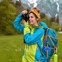 Vrouw met gele hoofdband en blauwgroene jas fotografeert in een groen, heuvelachtig landschap met rugzak.