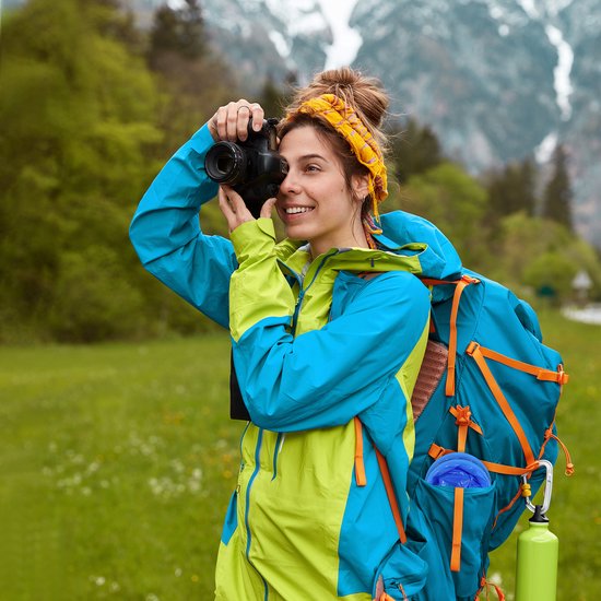 Vrouw met gele hoofdband en blauwgroene jas fotografeert in een groen, heuvelachtig landschap met rugzak.