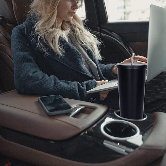 Zwarte thermobeker met rietje in de middenconsole van een auto, naast een telefoon en laptop.