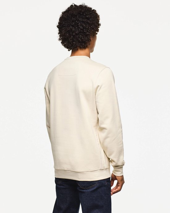 Man met krullend haar, van achteren gezien, draagt een beige trui met donkerblauwe jeans.