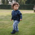 Jongetje met krullend haar in een donkerblauwe sweater en lichte jeans, draagt donkerblauwe hoge veterschoenen.