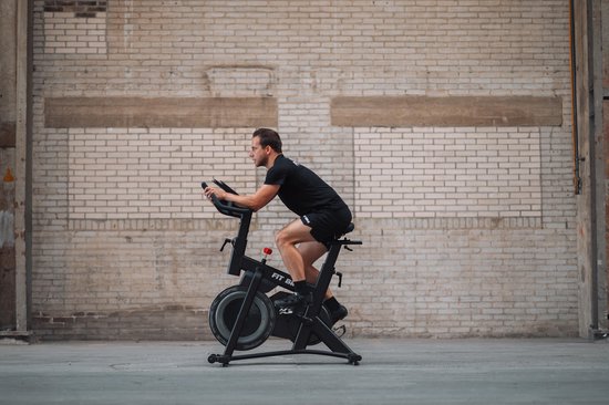 Zijaanzicht van een man die fietst op een zwarte spinfiets met een bakstenen muur op de achtergrond.