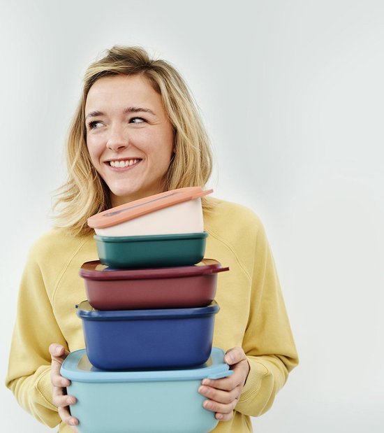 Une jeune femme souriante aux cheveux blonds porte un pull jaune et tient une pile de cinq boîtes de conservation colorées.