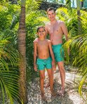 Jongens in turquoise zwembroeken met wit patroon, staand tussen palmbomen bij een zwembad.