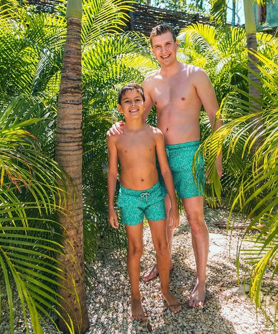Jongens in turquoise zwembroeken met wit patroon, staand tussen palmbomen bij een zwembad.