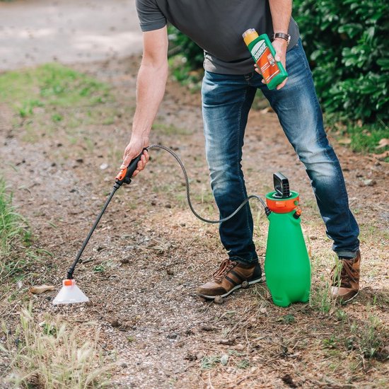 Man in spijkerbroek spuit met Roundup groen-oranje fles en spuitlans op de grond. Groene sproeier ernaast.
