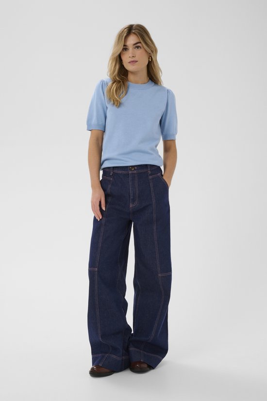 Een blonde vrouw in een lichtblauwe trui met korte, gepofte mouwen en een donkere spijkerbroek.