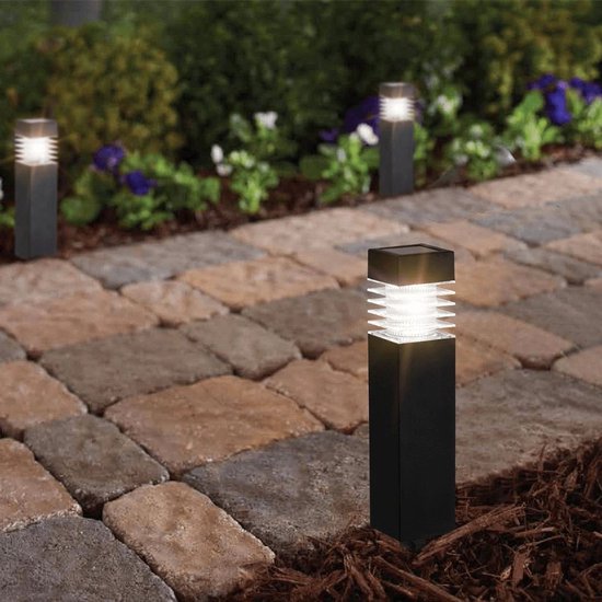 Zwarte vierkante tuinlamp met geribd glazen kap en warm wit licht, geplaatst in grind naast een terras.
