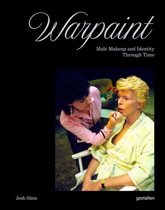 Een vrouw brengt make-up aan op David Bowie, die een geel geblokt ochtendjasje draagt. Titel: Warpaint. Auteur: Josh Sims.
