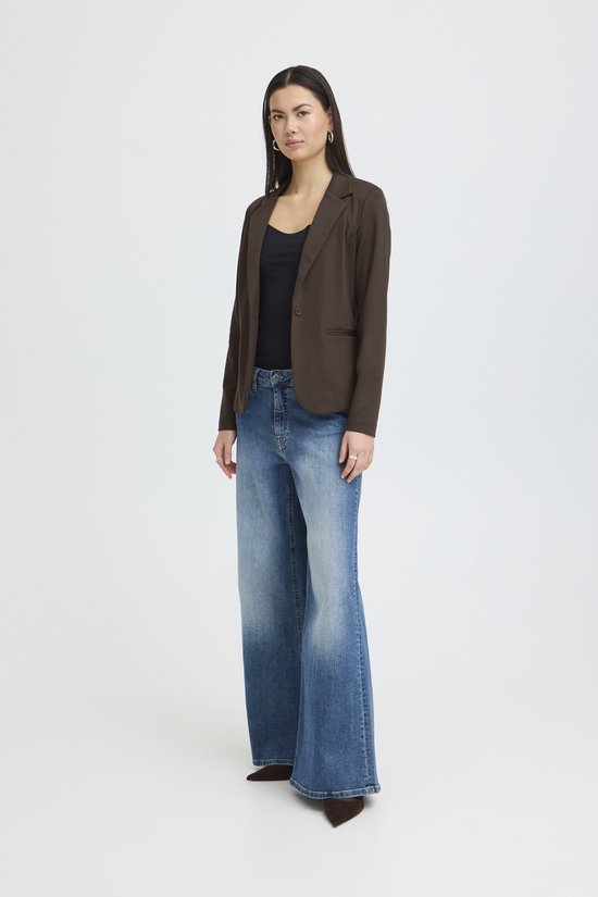 Vrouw, van de zijkant gezien, met lange donkere haren, draagt bruine blazer, zwart topje, wijde spijkerbroek.