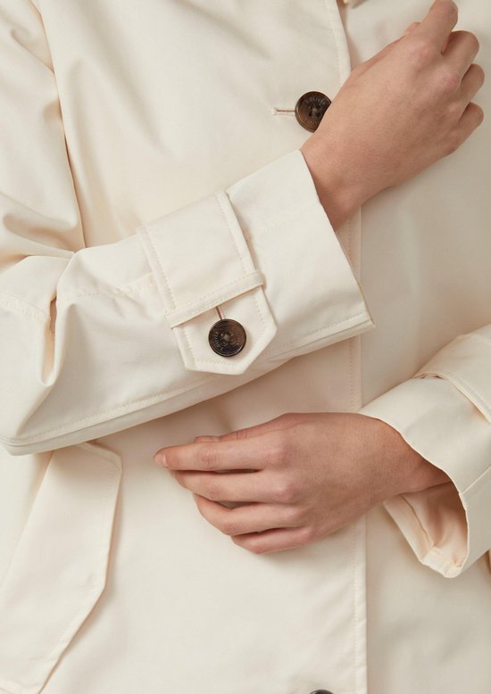 Crèmekleurige trenchcoat met bruine knopen, dichtbij de mouw met klep en knoop.
