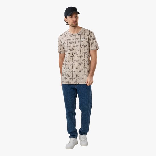 Man in vol ornaat, draagt beige T-shirt met palmbomenprint, spijkerbroek en zwarte pet.