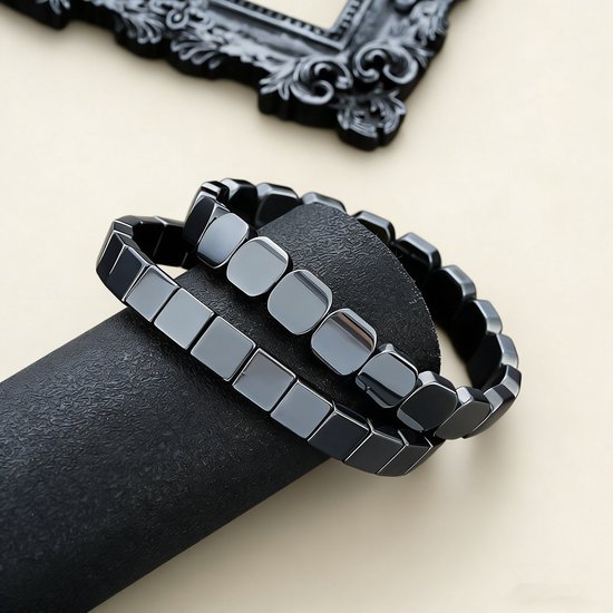 Twee zwarte, blokvormige armbanden van hematiet, een op een zwarte standaard.