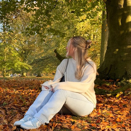 Blonde vrouw in beige trui en lichte jeans, zittend op herfstbladeren bij een boom.