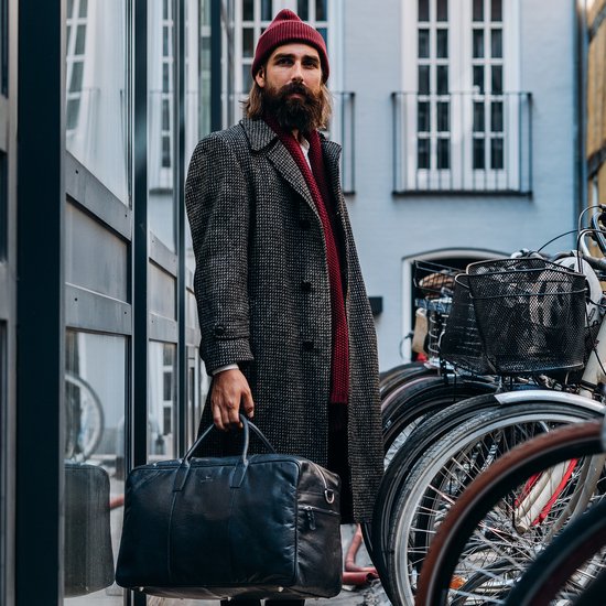 Man met baard draagt een wijnrode muts, sjaal, grijze jas en draagt een zwarte tas.