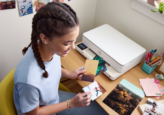 Vrolijke jonge vrouw met vlecht met clipjes, selecteert foto's bij een witte HP Envy printer.
