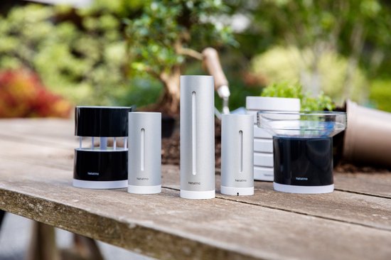 Diverse Netatmo weerstation modules op een houten tafel in de tuin, waaronder een regenmeter, thermometer en anemometer.