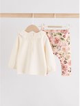 Roomwitte longsleeve met rucheskraag en een legging met bloemenprint, hangend aan een kledingrek.