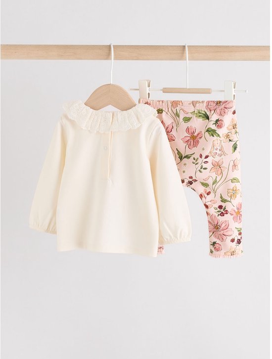Roomwitte longsleeve met rucheskraag en een legging met bloemenprint, hangend aan een kledingrek.