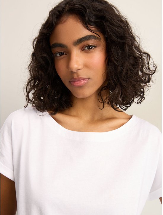 Een vrouw met krullend donker haar draagt een wit T-shirt met opgerolde korte mouwen en een ronde hals.