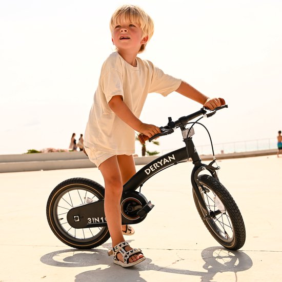 Petit garçon aux cheveux blonds sur un vélo Deryan noir, regardant au loin. Assis sur une place ensoleillée avec des gens à l'arrière-plan.