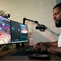 Homme avec un casque jouant à des jeux vidéo derrière deux écrans d'ordinateur, avec une webcam et un microphone.