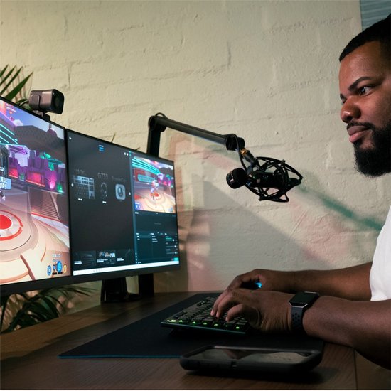 Homme avec un casque jouant à des jeux vidéo derrière deux écrans d'ordinateur, avec une webcam et un microphone.