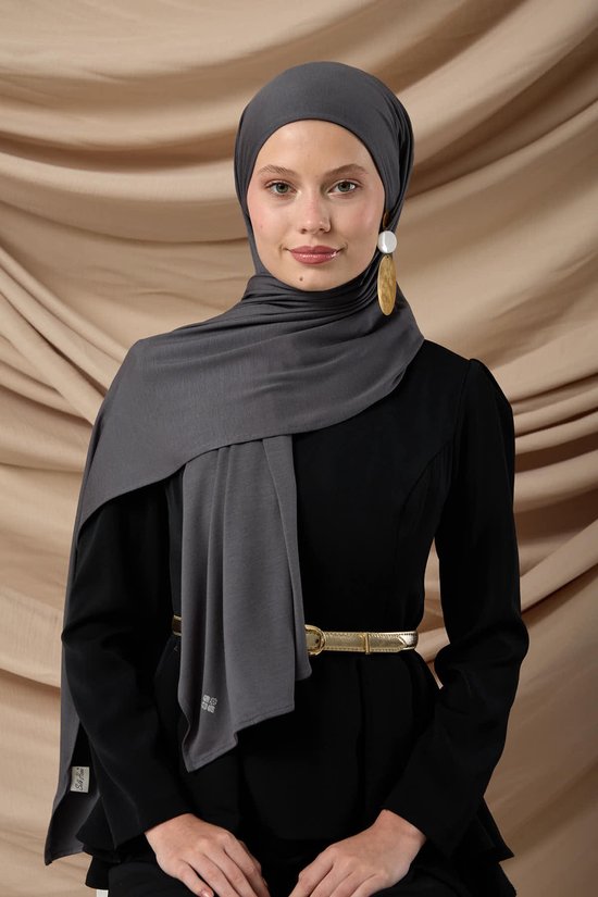 Model met donkergrijze jersey hoofddoek, die over een zwarte jurk is gedrapeerd. De hoofddoek valt soepel en heeft een luxe uitstraling.