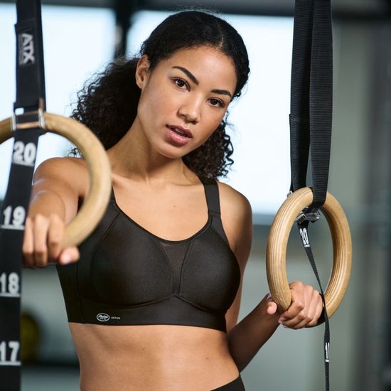 Een vrouw met krullend haar hangt aan sportringen, draagt een zwarte sportbeha.