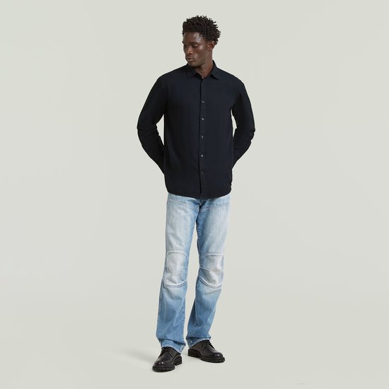 Man, hele lichaam, kijkt weg, draagt een donkerblauw regular fit overhemd, handen achter de rug.