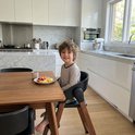 Enfant riant dans une chaise haute Deryan noire avec pieds aspect bois, assis à une table en bois.