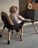 Een klein kind met krullend haar leest een boek zittend aan een zwarte tafel met houten poten. De stoel is bruin.