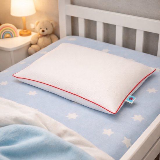 Wit kussen met rode bies op een kinderbed met een blauw sterren dekbedovertrek. Naast het bed staat een nachtkastje met een lampje en een teddybeer.