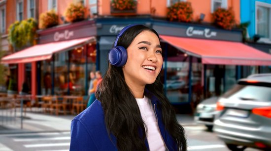 Vrolijke vrouw met lange zwarte haren draagt een marineblauwe jas en een donkerblauwe JBL koptelefoon in een straat.
